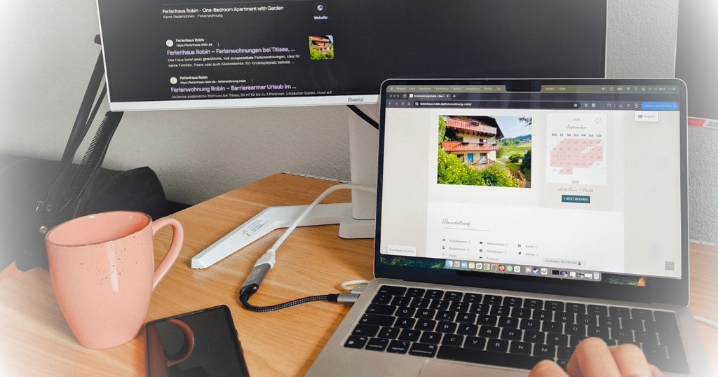 Laptop mit der Website einer Ferienwohnung samt Verfügbarkeitskalender und Buchungsbutton, daneben ein Monitor mit den Google-Suchergebnissen zum Objekt